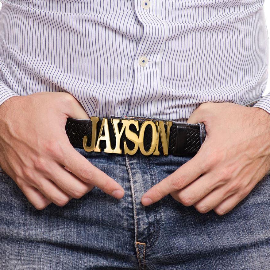 Man in striped shirt pointing to a black belt with custom gold buckle spelling 'JAYSON' against a denim jeans background.