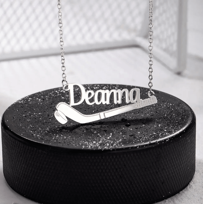 A silver necklace with the name "Deanna" elegantly scripted, featuring a hockey stick and puck design, displayed on a black, glittery circular platform.