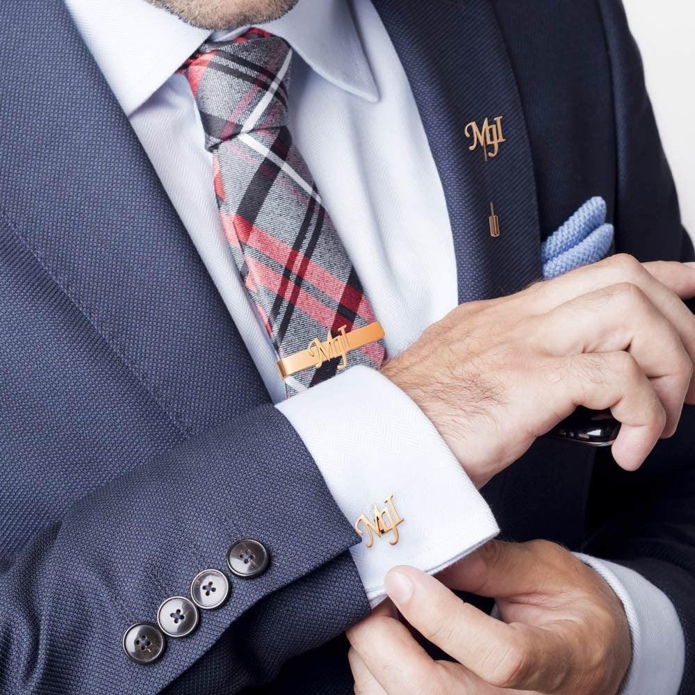 A man in a navy suit adjusting rose gold cufflinks and a tie bar with the monogram "MJT" on his shirt cuff and lapel.