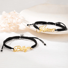 Two black braided bracelets with gold name tags reading "Billy" and "Karen," adorned with heart shapes, displayed on a white surface next to delicate flowers.