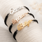 Three black cord bracelets with nameplates in different metals: "Emma" in silver, "Sophia" in rose gold, and "Billy" in gold, each with a small heart at the end.