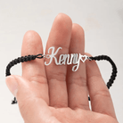 A black cord bracelet with a silver nameplate reading "Kenny," held between two fingers, featuring a small heart at the end of the name.