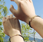 Two hands wearing black cord bracelets with gold nameplates reading "Klyen" and "Billy," featuring small hearts at the end of the names, with a clear blue sky background.