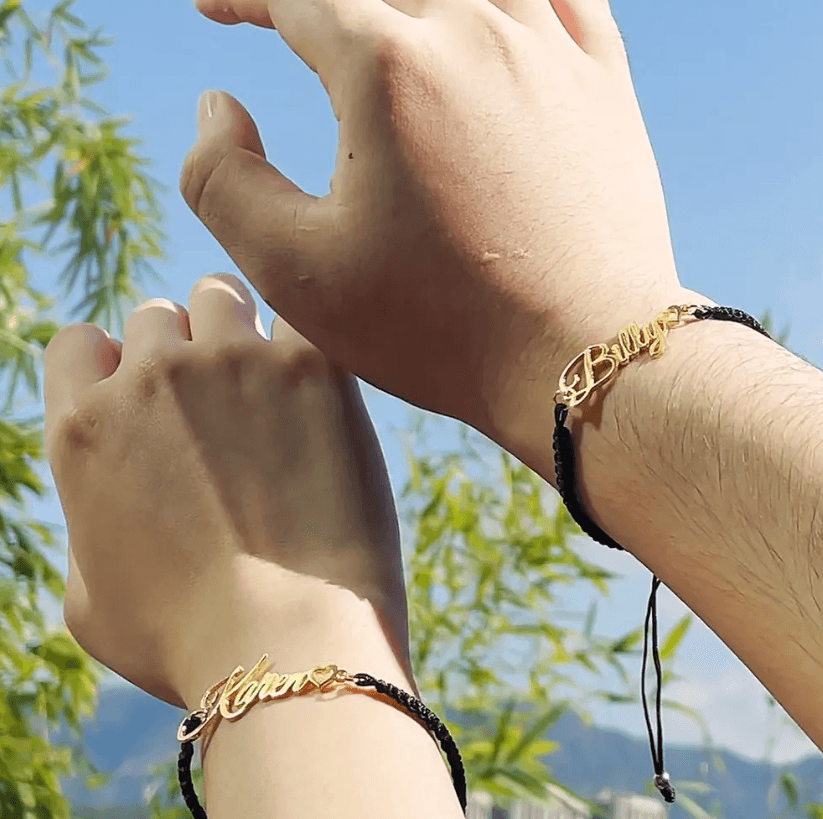 Two hands wearing black cord bracelets with gold nameplates reading "Klyen" and "Billy," featuring small hearts at the end of the names, with a clear blue sky background.