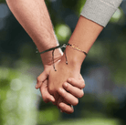 Two hands holding each other, each wearing a black cord bracelet with a gold nameplate. One bracelet reads "Karen," and both have small hearts at the end of the names.