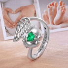 Silver memorial ring with angel wings design and a green heart-shaped stone nestled between baby feet, with the inscription "Forever in my heart" and a background of photos showing heart-shaped hands on a pregnant belly and baby feet.