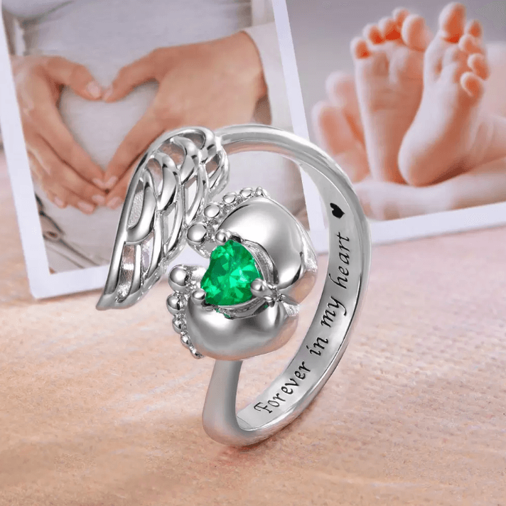 Silver memorial ring with angel wings design and a green heart-shaped stone nestled between baby feet, with the inscription "Forever in my heart" and a background of photos showing heart-shaped hands on a pregnant belly and baby feet.