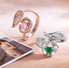 Two memorial rings with angel wings and heart-shaped stones cradled by feet; one rose gold with pink stone and "Sophia 02.10.2021" engraving, one silver with green stone and "Forever in my heart" inscription, beside a photo of a pregnant woman.