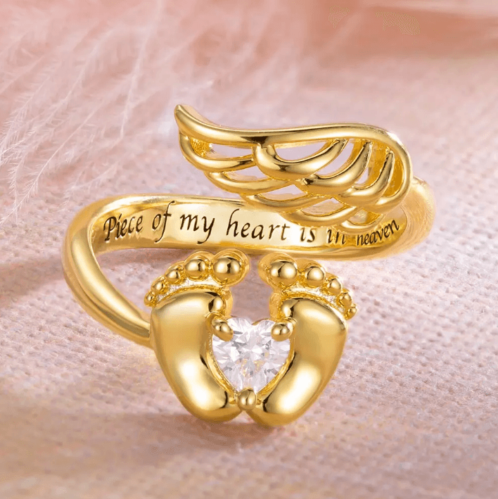 A gold-toned memorial ring with a heart-shaped stone cradled by feet, angel wings on top, and an inscription "Piece of my heart is in heaven".