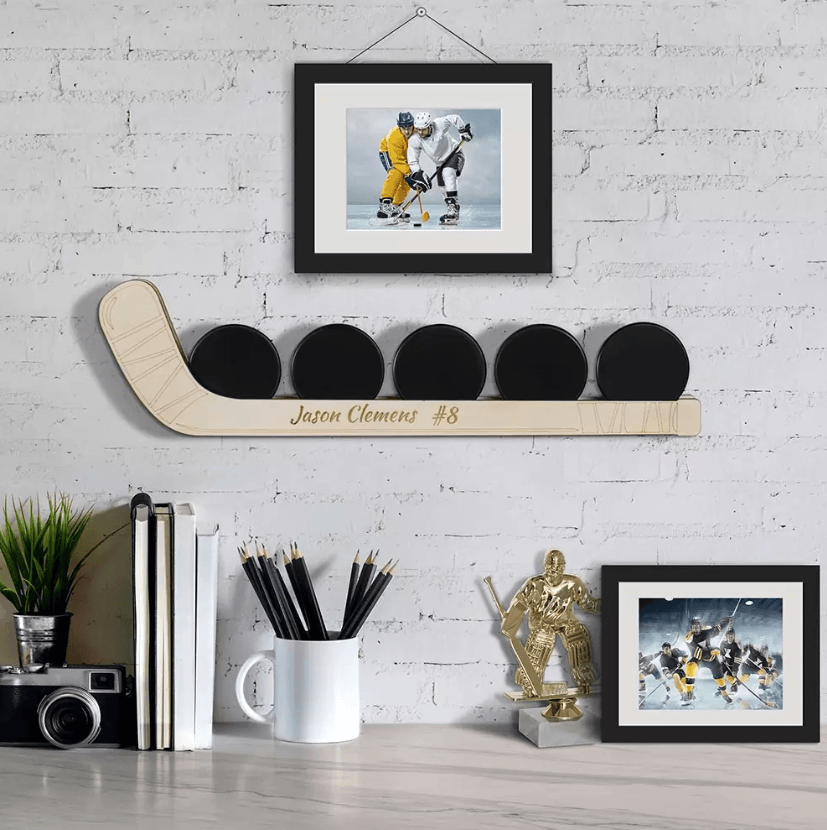 Customizable wooden hockey puck display shelf with five pucks, personalized with the name "Jason Clemens #8", shown mounted on a wall above a desk with books, pencils, and a hockey trophy.