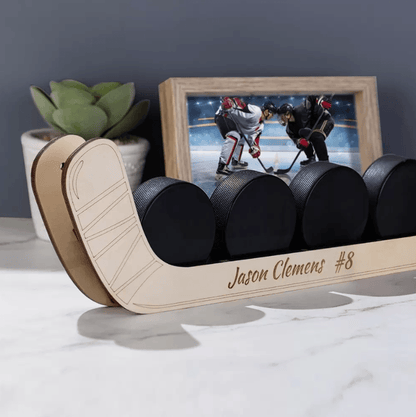Close-up of a customizable wooden hockey puck display shelf with five pucks, personalized with the name "Jason Clemens #8", placed on a table with a plant and a framed hockey photo in the background.