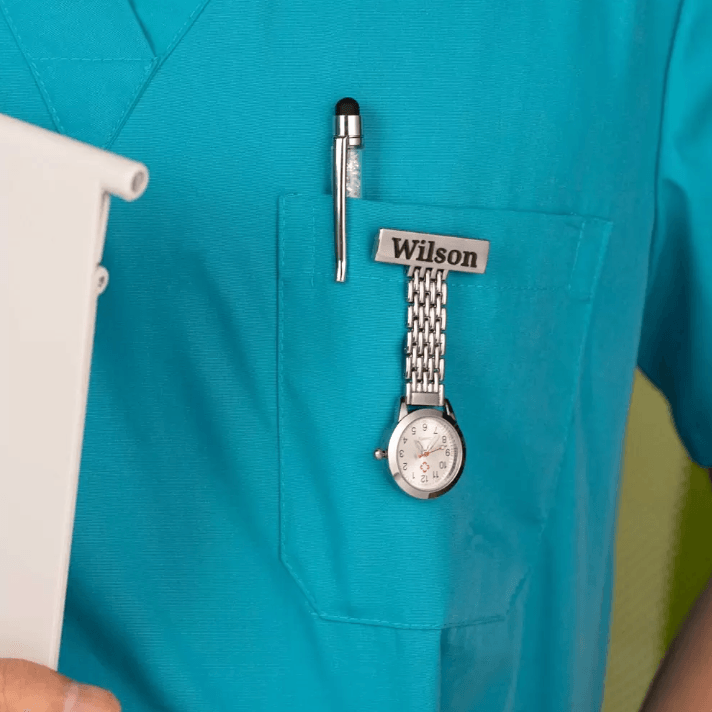 A healthcare professional in turquoise scrubs with a pen, a name badge reading "Wilson," and a silver watch clipped to the chest pocket.