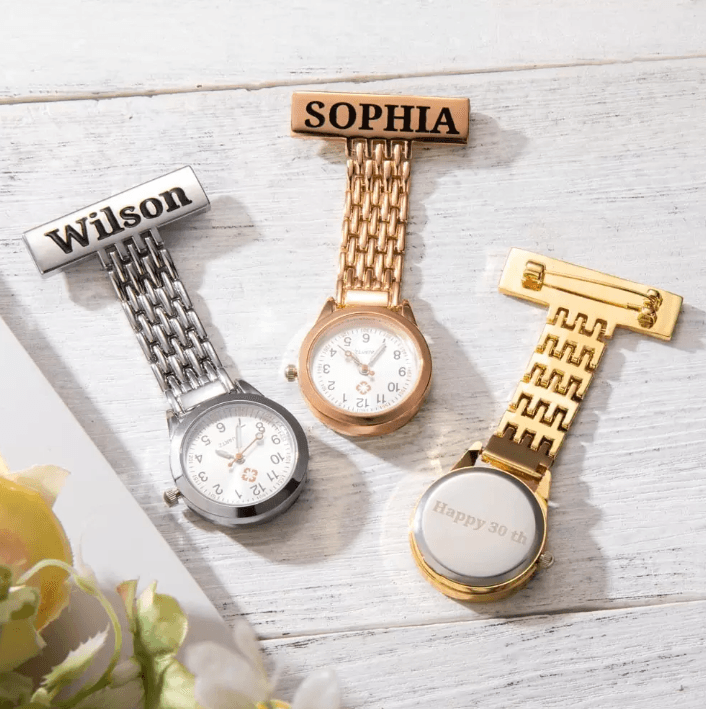 Three nurse watches on a wooden surface: a silver one labeled "Wilson," a rose gold one labeled "Sophia," and a gold one engraved with "Happy 30th" on the back.