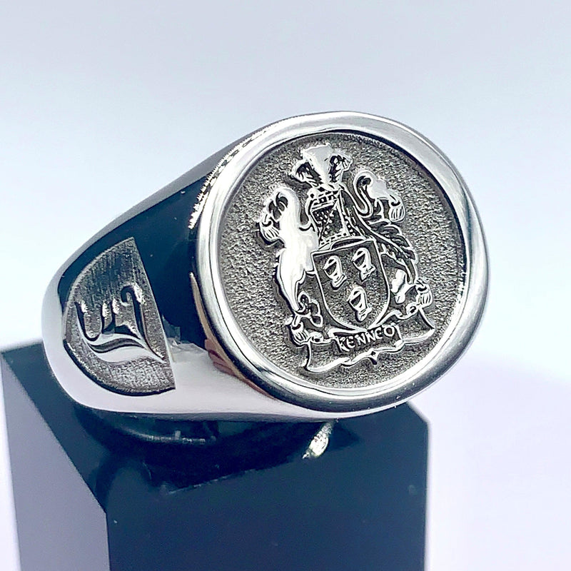A silver signet ring with the Kennedy family crest and an Old English 'J' initial on the side, displayed on a black stand.