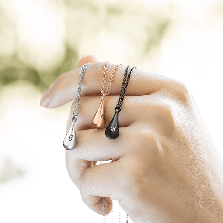 Hand holding three teardrop pendant necklaces in silver, rose gold, and black.