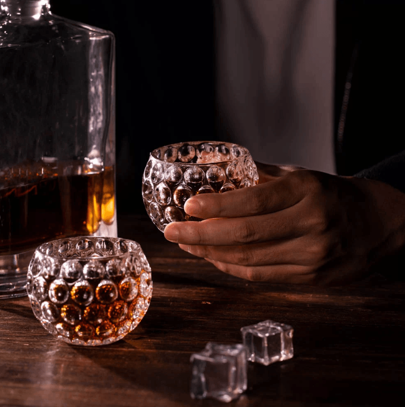 Person holding a golf ball-shaped whiskey glass, with another glass and a decanter filled with whiskey on a wooden table, illuminated warmly.