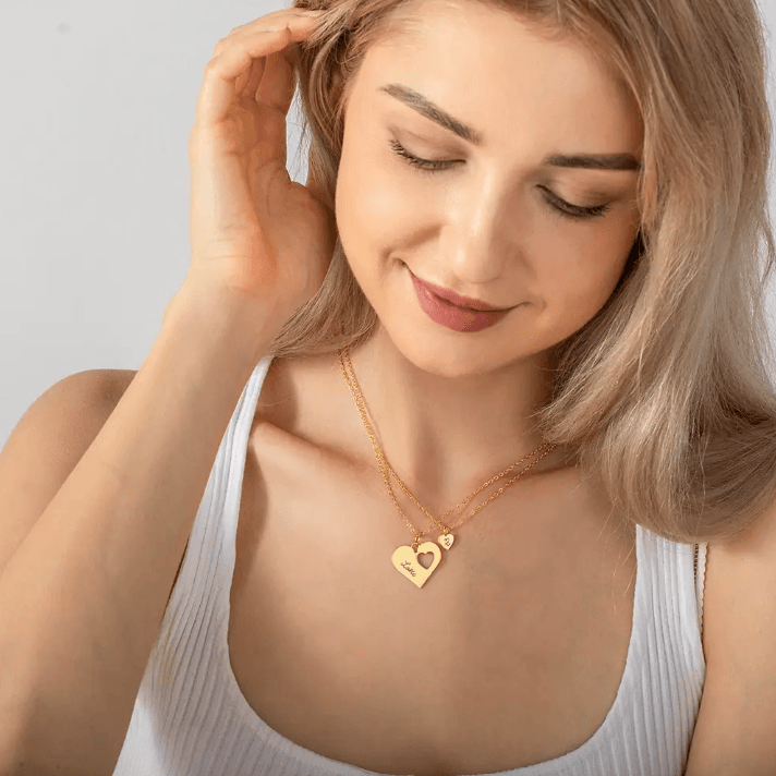 Smiling woman wearing a layered gold heart necklace set with engravings 'Lotte' and 'R', highlighting the elegant and personalized jewelry design against a white top.