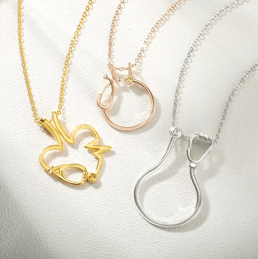 Gold, rose gold, and silver ring holder necklaces with heart, stethoscope, and EKG designs. Each pendant features a customizable birthstone, ideal for medical professionals.