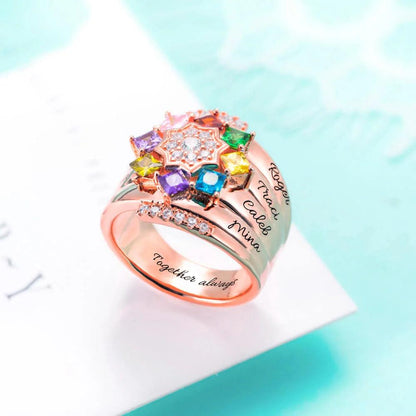 A rose gold ring featuring a star-shaped arrangement of colorful gemstones, engraved with multiple names and small diamonds on its band, placed on a light blue background.