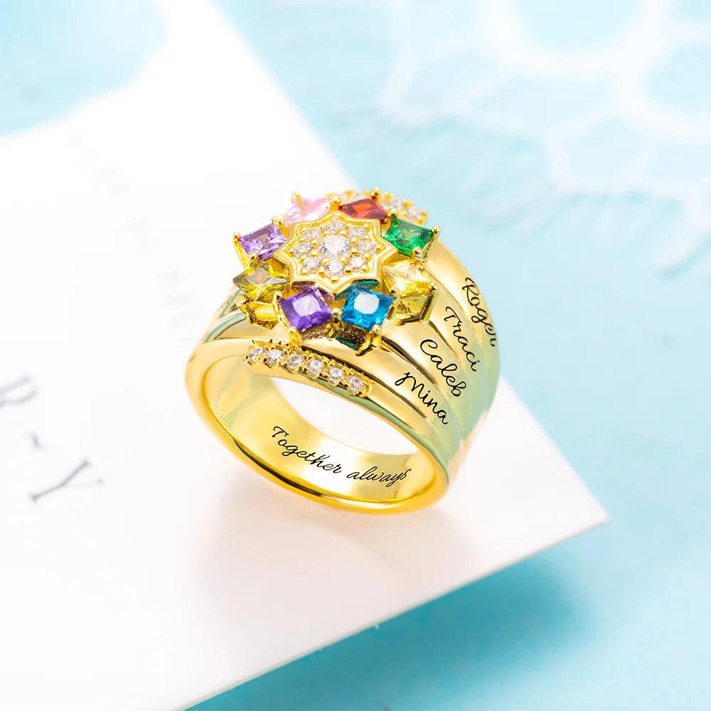 A gold ring adorned with colorful gemstones arranged in a star shape, featuring engraved names on its band and small diamonds on the sides, placed on a light blue background.