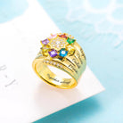 A gold ring adorned with colorful gemstones arranged in a star shape, featuring engraved names on its band and small diamonds on the sides, placed on a light blue background.