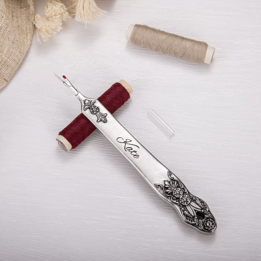 Personalized seam ripper with 'Kate' engraving on silver floral handle, red thread spool, and beige spool on a white surface.