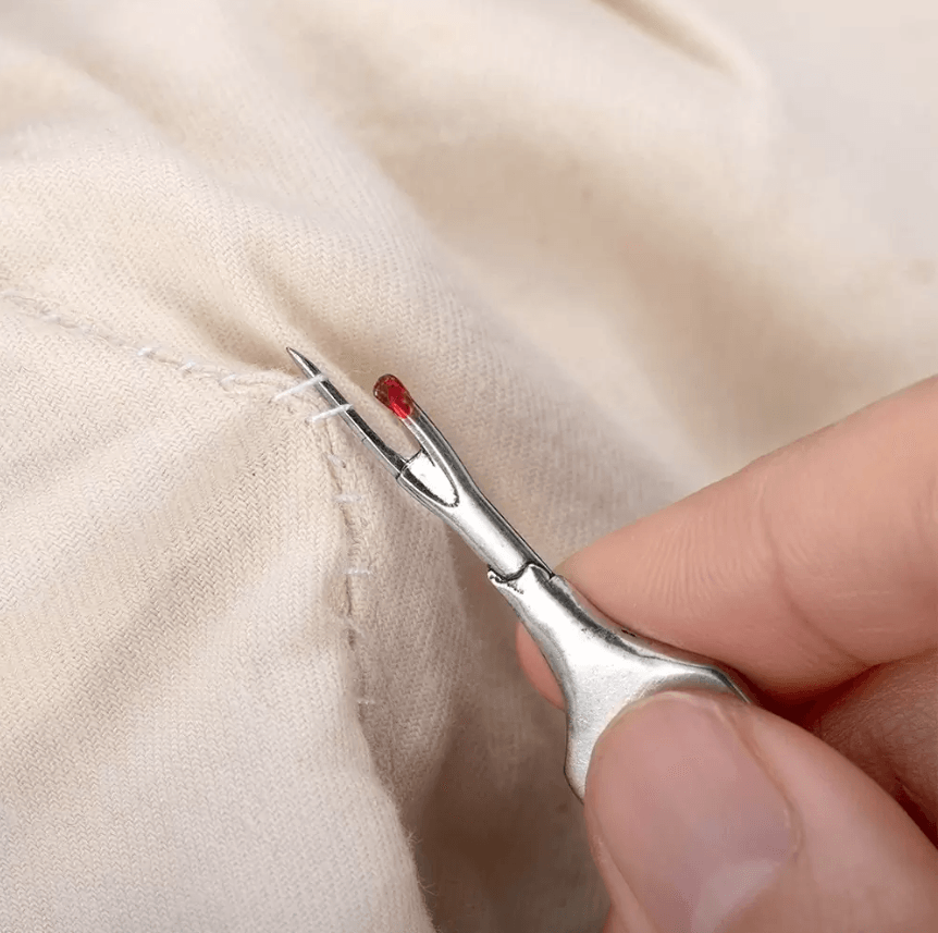 Close-up of a hand using a silver seam ripper with a red tip to remove stitches from beige fabric.