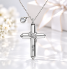 A silver cross cremation urn pendant necklace with a small round clear gemstone and a delicate silver wing detail, shown on a reflective white surface with soft blurred white flowers and a gift box in the background.