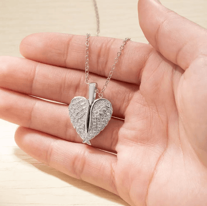 Hand holding a silver angel wing heart locket with intricate crystal details on a chain.