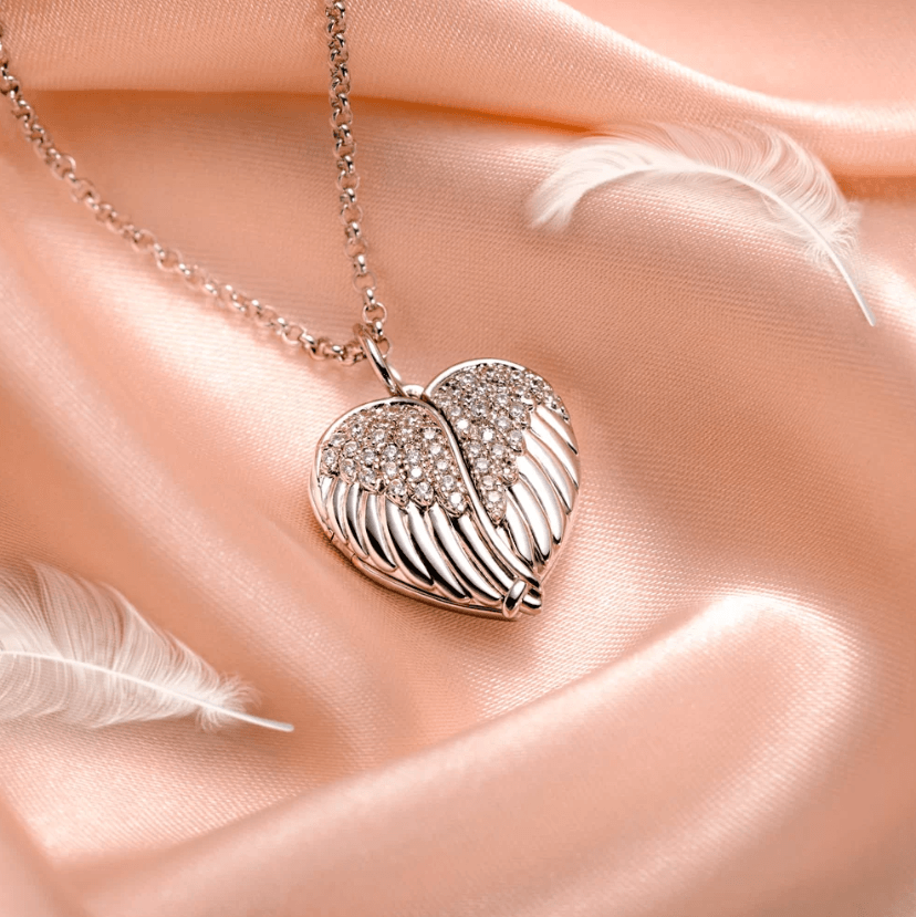 A silver heart-shaped pendant adorned with sparkling crystals, designed to resemble angel wings, rests elegantly on a soft pink satin fabric with delicate white feathers.