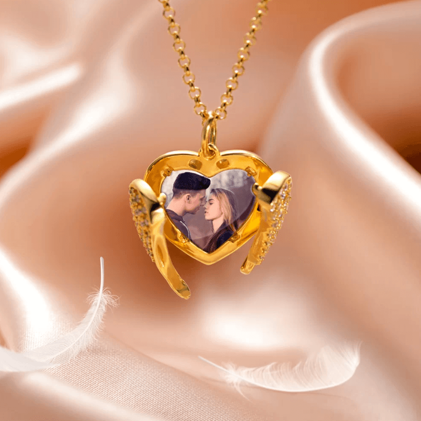 A golden heart-shaped locket with angel wing details opens to reveal a photo of a couple touching foreheads, resting on soft pink satin fabric with delicate white feathers.