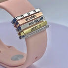 Pink Apple Watch band with personalized gold, silver, and diamond-like engraved charms featuring the names "Belbren," "Avelina," and "Jacky."