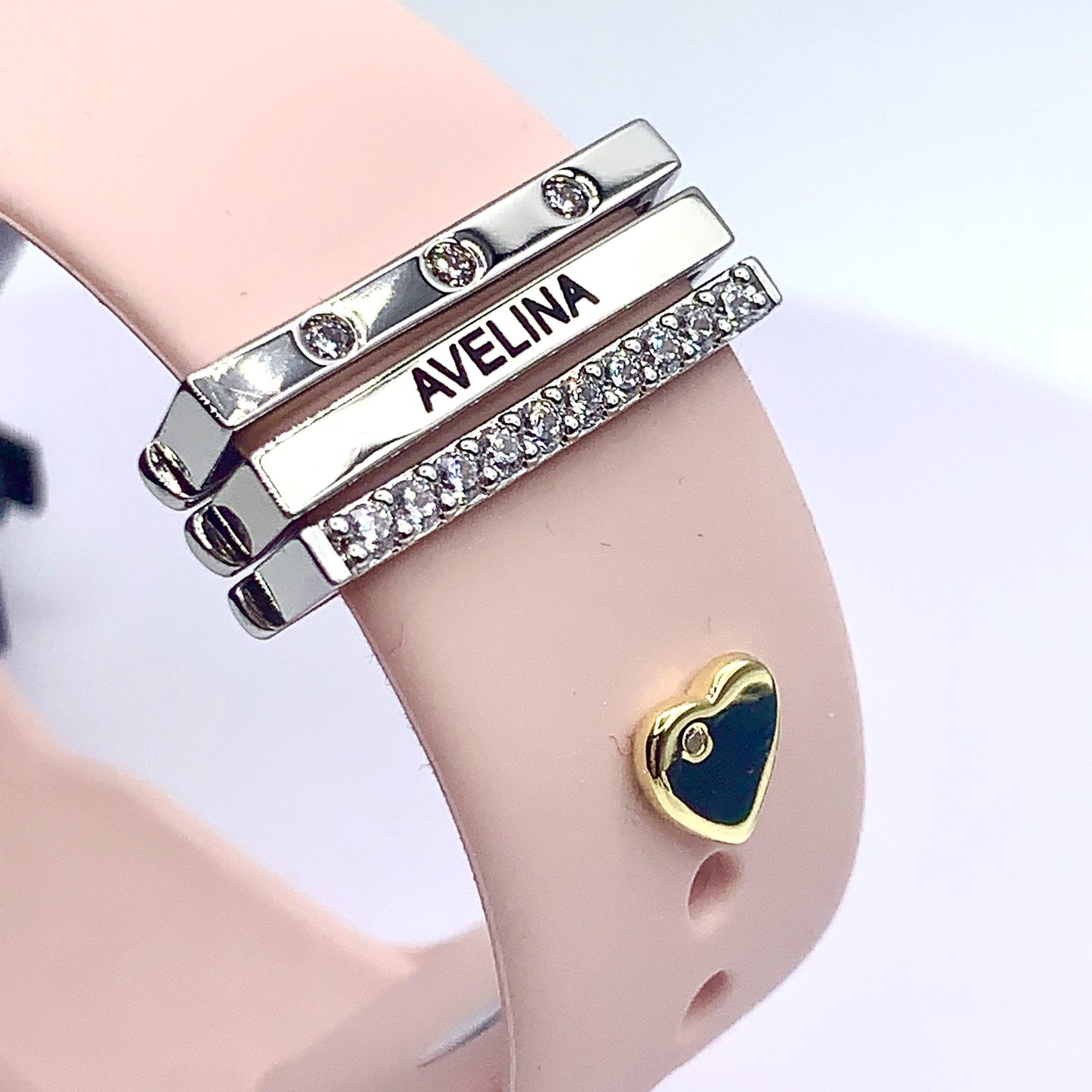  Close-up of a pink Apple Watch band with personalized silver and diamond-like engraved charms featuring the name "Avelina" and a gold heart accent.