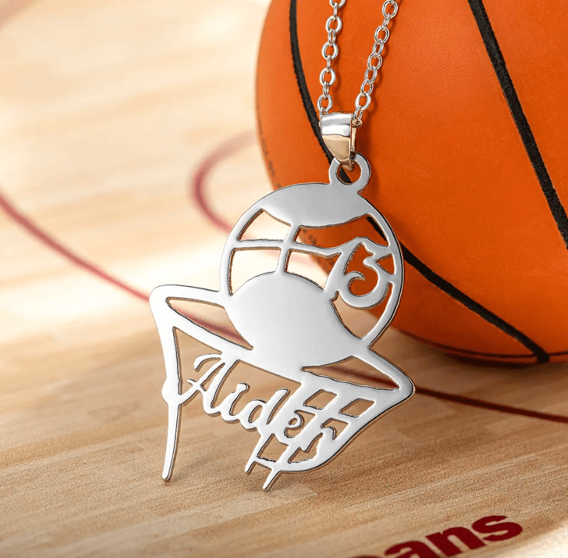 Silver basketball pendant necklace with customized name and jersey number '15', displayed near a basketball on a wooden court.