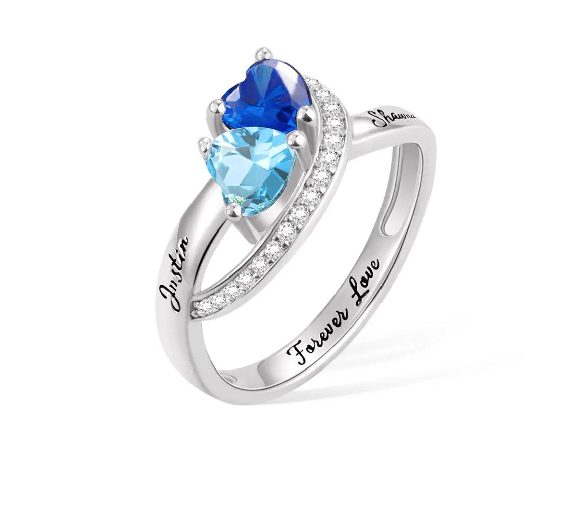 A silver ring with a heart-shaped blue gem, flanked by clear crystals, inscribed with "Justin" and "Shawna Forever Love