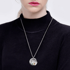 A woman wearing a black turtleneck sweater and a silver round locket necklace with a tree design and colorful gemstones.