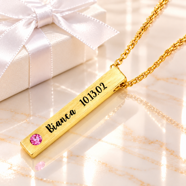 Personalized gold 4-sided bar necklace with brushed finish, custom Bianca name and date engraving, and pink birthstone, displayed on glossy cream marble beside a white gift box with satin ribbon.