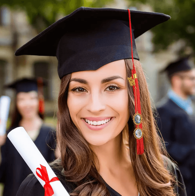 A young woman in a graduation cap and gown smiles while holding her diploma, with a red tassel adorned with gold medallions on her graduation cap. Other graduates are visible in the background.