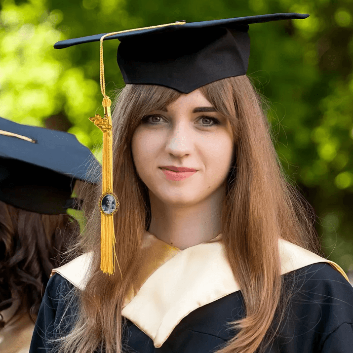 A young woman in a graduation cap and gown, with a gold tassel adorned with an ornate frame holding a photo, stands outdoors with another graduate in the background.