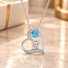 Silver Highland cow heart necklace with blue birthstone and sparkling crystal accents, displayed on a soft ivory marble background.