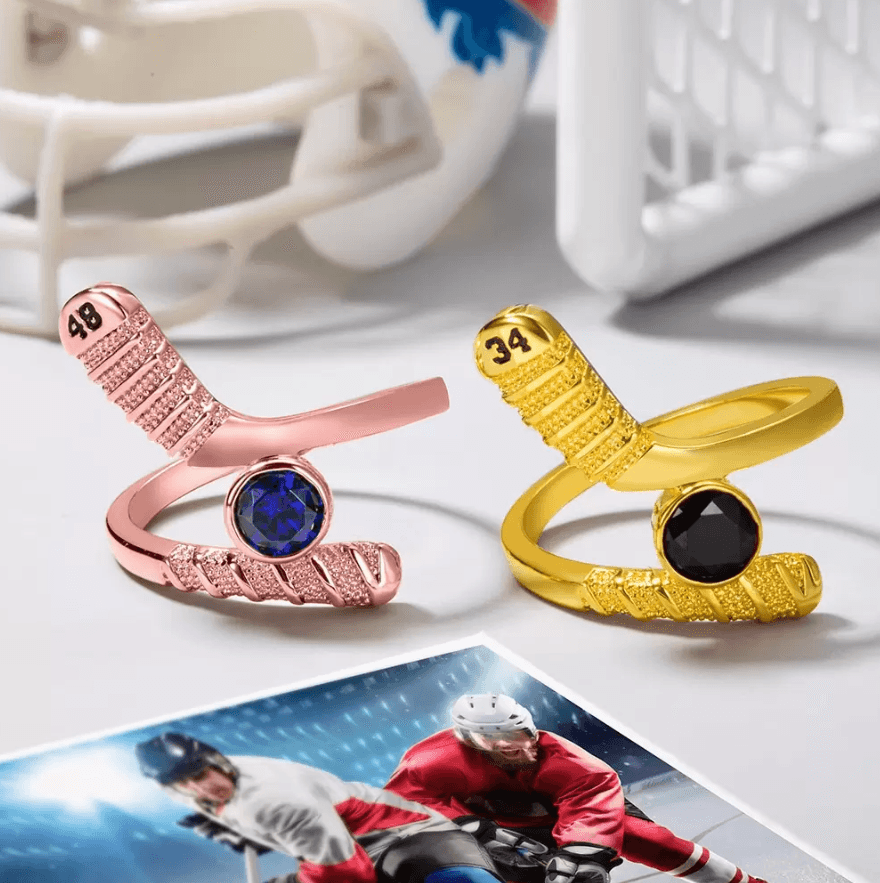 Rose gold and yellow gold hockey stick-shaped rings with blue and black gemstones, numbers '48' and '34', showcased near hockey gear.