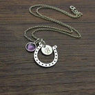 Personalized silver horseshoe necklace with engraved initial disc and purple birthstone charm, symbolizing luck and protection, displayed on a wooden surface.