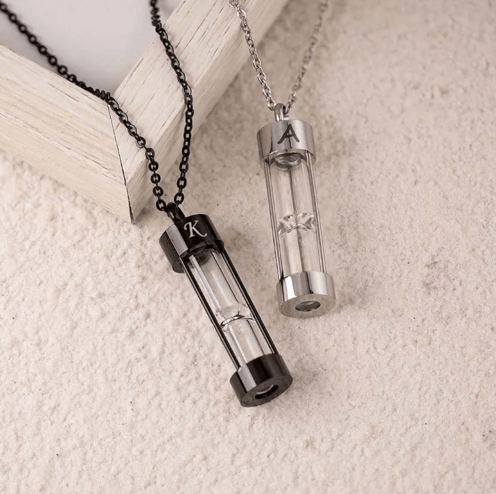 Two hourglass urn necklaces with initials 'K' and 'A' in black and silver, resting on a textured background.