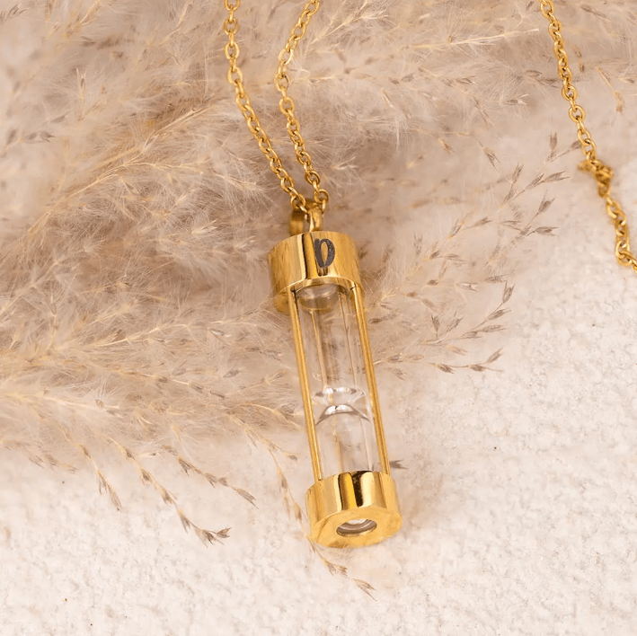 Gold hourglass urn necklace with personalized initial 'D', displayed on a fluffy, soft background.