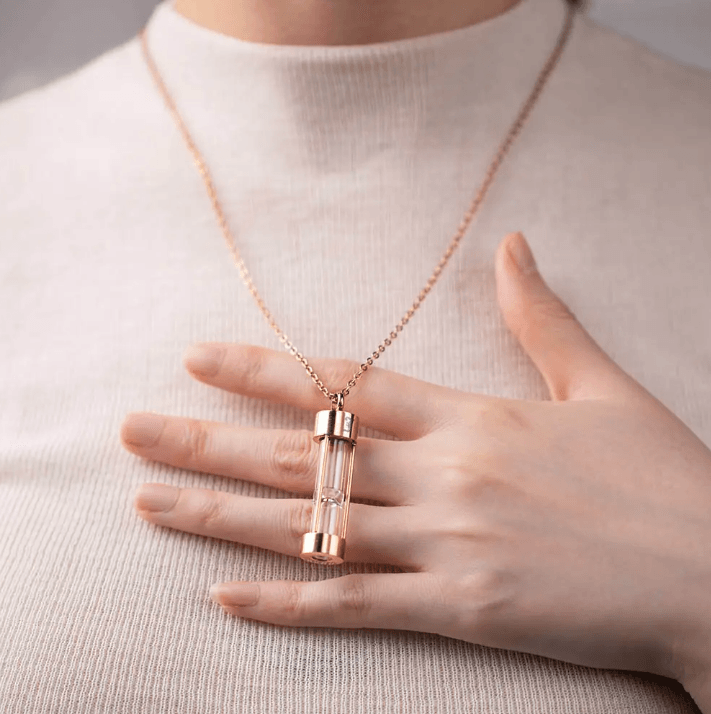Woman wearing a rose gold hourglass urn necklace, holding it gently with a soft sweater background.