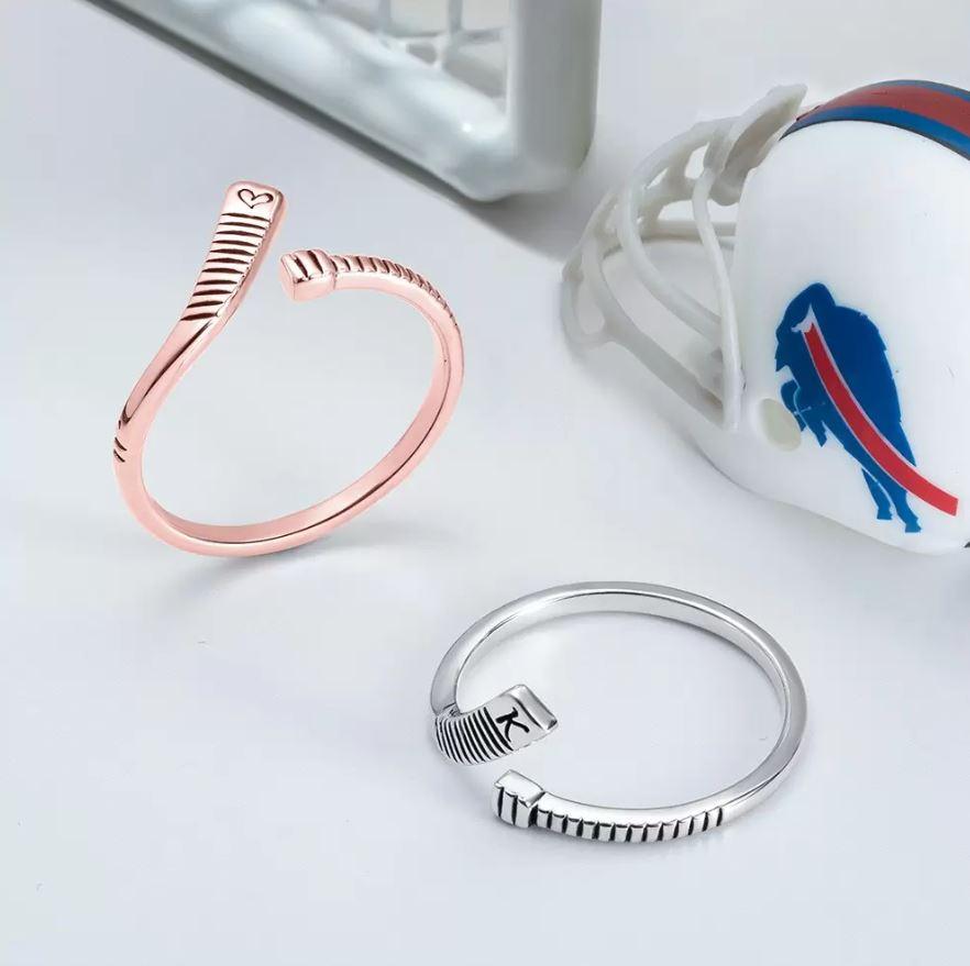 Two customizable hockey stick-shaped rings in rose gold and silver, featuring a heart icon and the letter "K," placed near a hockey helmet.