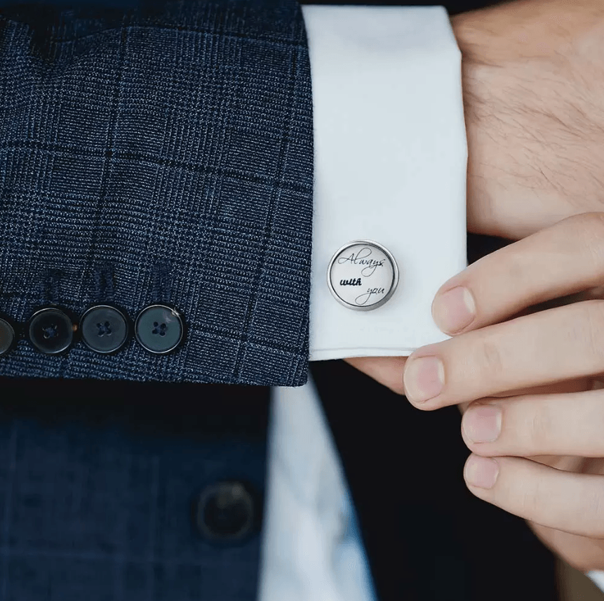 Close-up of a man's hand adjusting a white shirt cuff with a silver cufflink engraved with 'Always with you'.