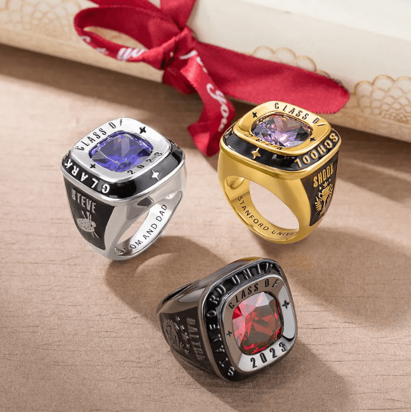 Three year class rings with colorful gemstones and engraved details, displayed on a surface with a red ribboned scroll in the background.