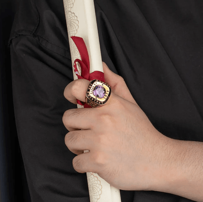 A person wearing a gold class ring with a purple gemstone, holding a rolled diploma tied with a red ribbon, dressed in black graduation attire.