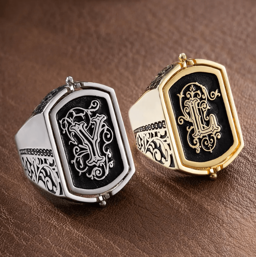 Two ornate men's flip rings, one silver and one gold, each with black inlay and embossed double initials, on a leather background.